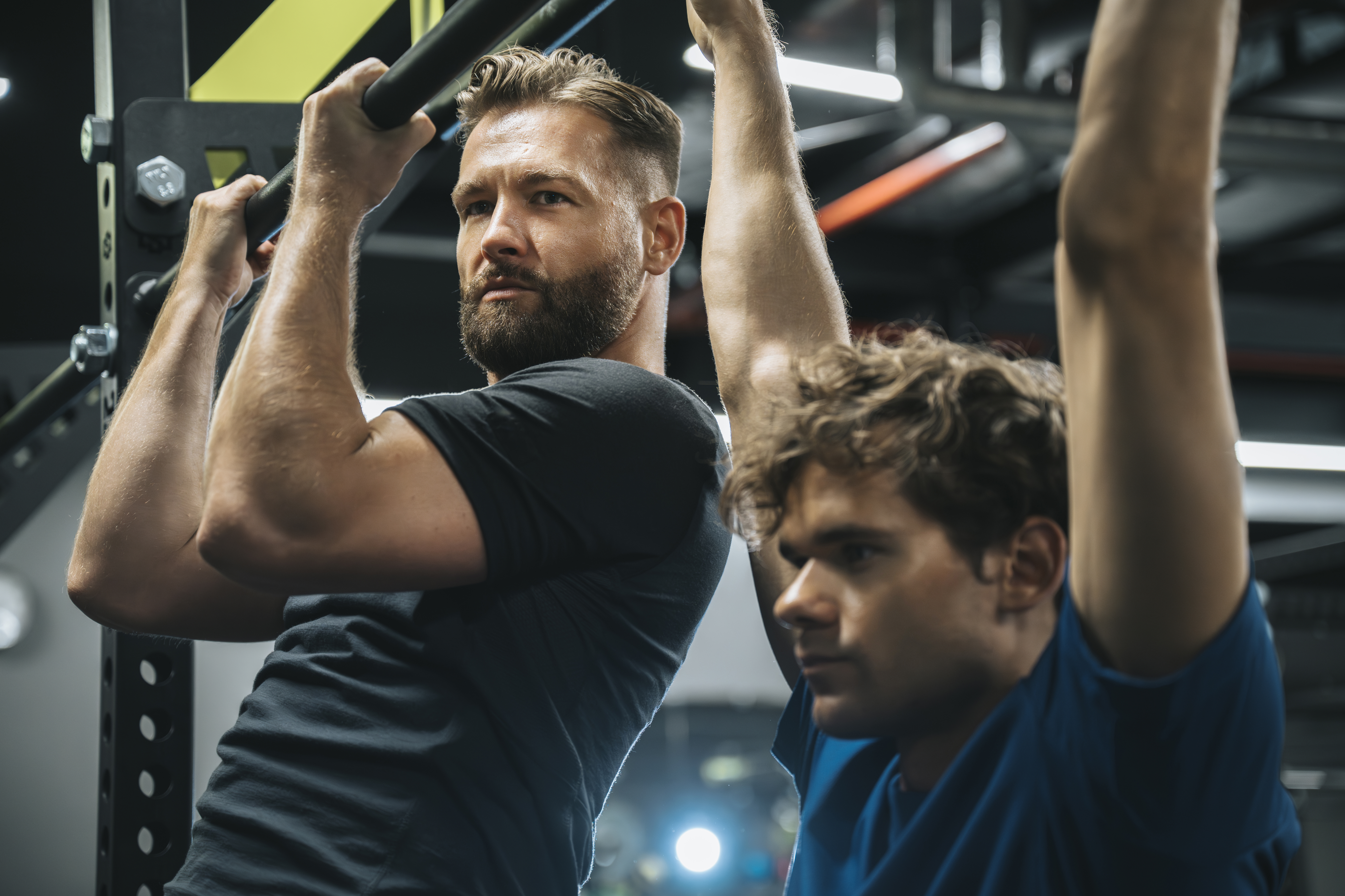 Dwóch mężczyzn trenuje na drążku w siłowni, wykonując ćwiczenia siłowe na podciąganie.