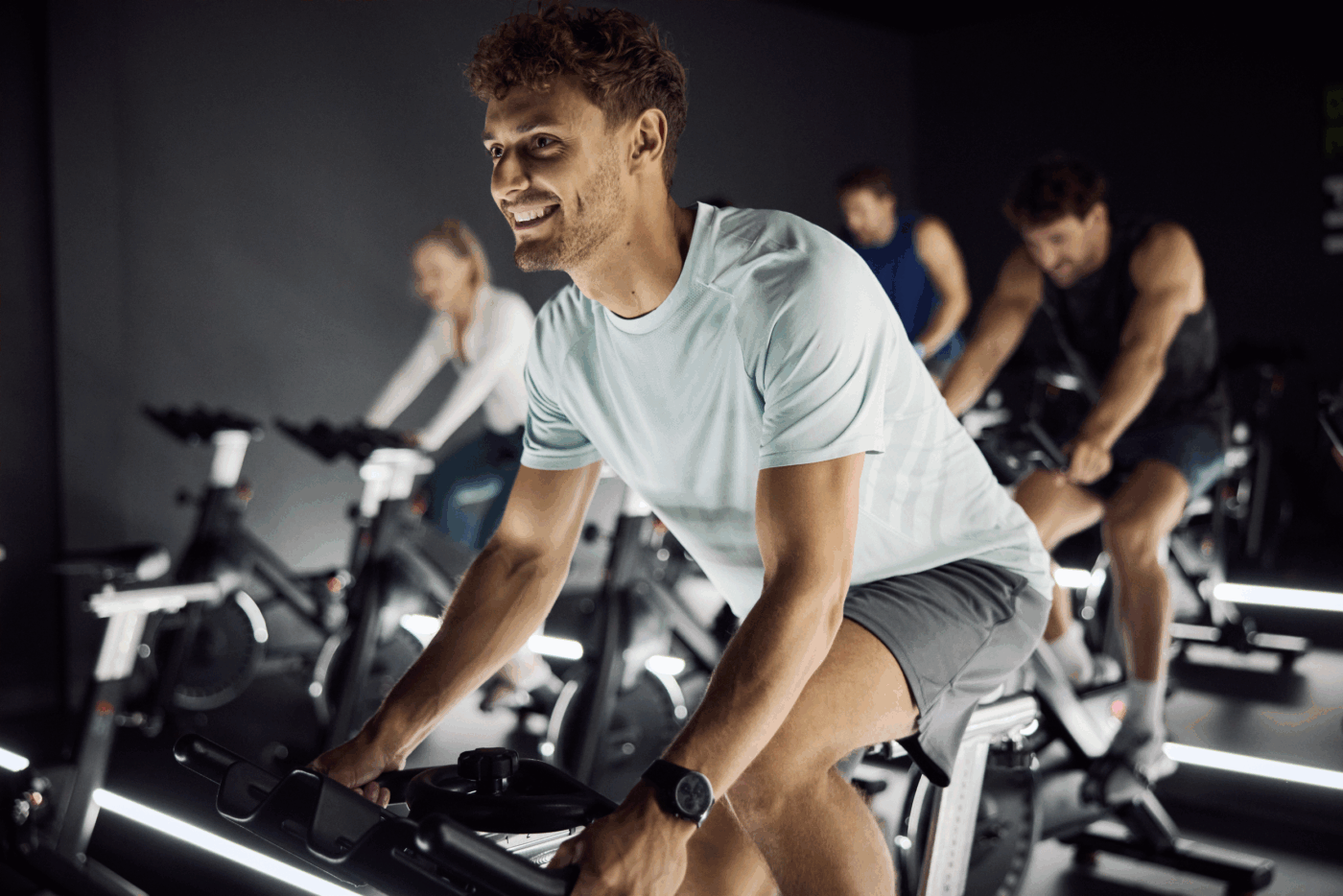 Учасники класу Indoor Cycling у фітнес-клубі тренуються на велотренажерах, усміхнений чоловік на передньому плані.