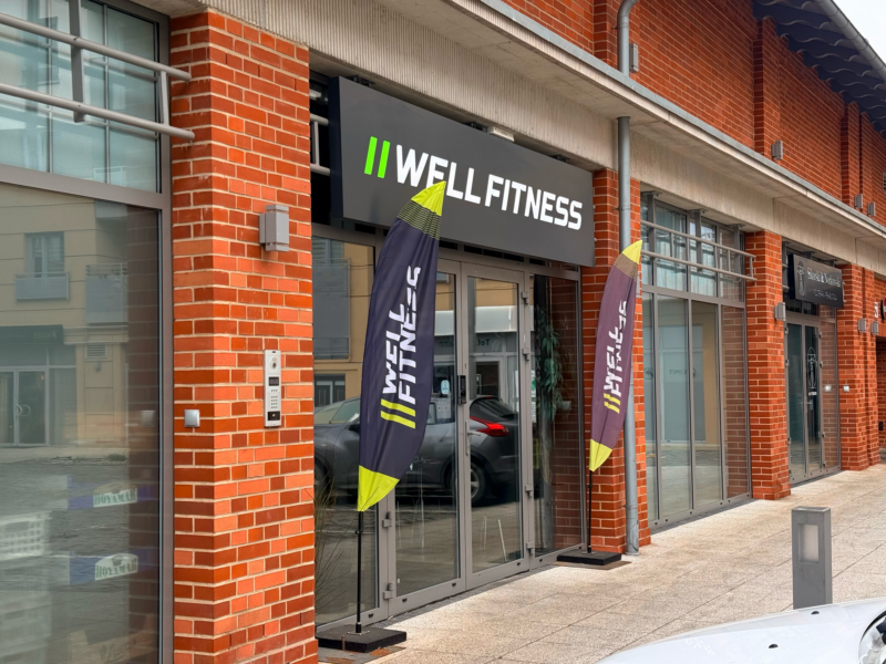 Вивіска клубу «Well Fitness» розміщена над заскленим входом до будівлі з червоної цегли; біля дверей видно два вертикальні рекламні банери.