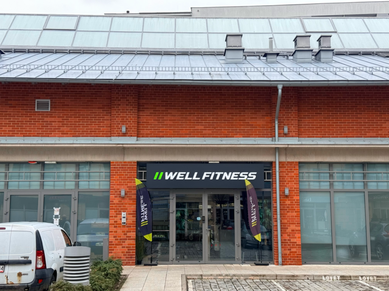 Фасадна стіна клубу Well Fitness у Вроцлаві на вулиці Тенчовій, з чітким логотипом над входом та двома рекламними банерами по боках дверей.