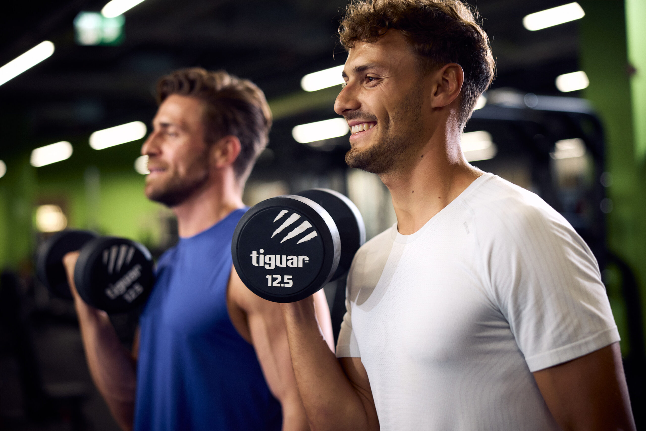 Двоє чоловіків тренуються з гантелями у спортзалі, виконуючи вправи для зміцнення рук.