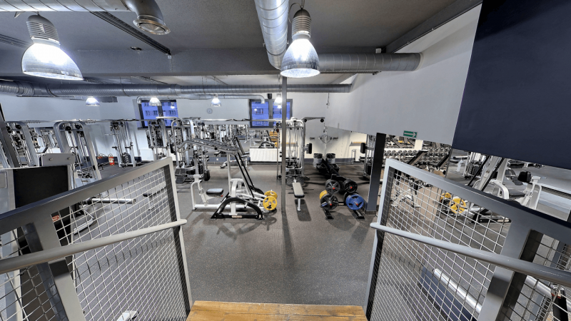 Zdjęcie zrobione z podestu, z widokiem na salę z maszynami do ćwiczeń siłowych.