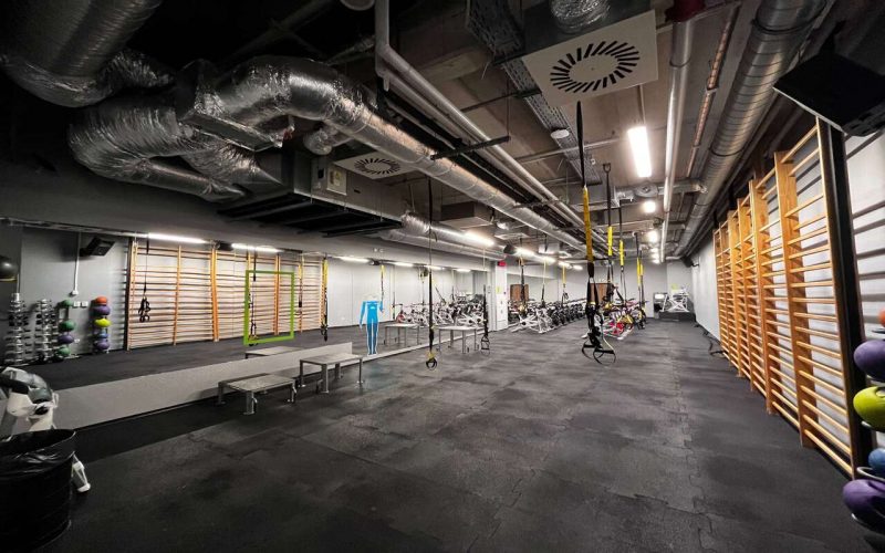 Sala fitness z lustrem. Z sufitu zwisają taśmy do ćwiczeń TRX. Pod ścianą zamontowane są drabinki oraz stoją stojaki z akesoriami do ćwiczeń.
