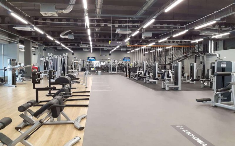 Duża sala z maszynami do ćwiczeń poszczególnych partii mięśni. W tle strefa do ćwiczeń ogólnorozwojowych oraz drążki.