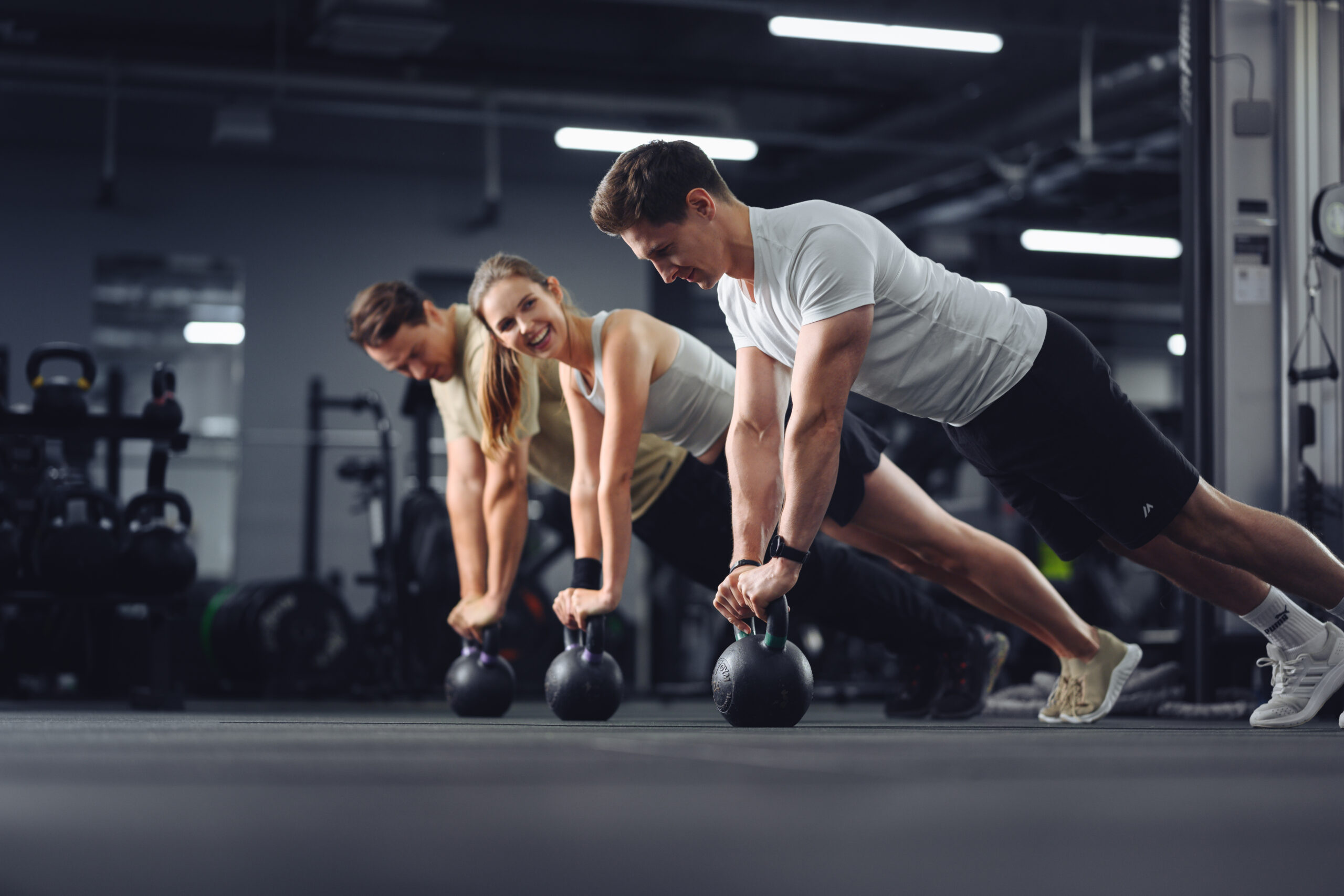 Grupa osób ćwiczących w siłowni – trzy osoby wykonują podpór na kettlach podczas zajęć funkcjonalnych, uśmiechnięte i skoncentrowane na treningu.