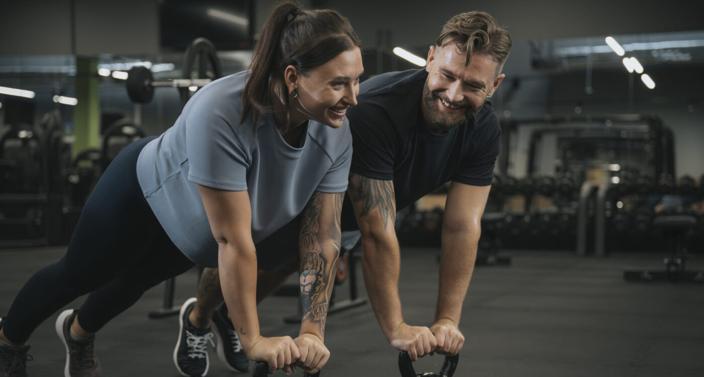 Trener i uczestniczka zajęć wykonujący deskę na kettlebellach w siłowni, uśmiechając się do siebie