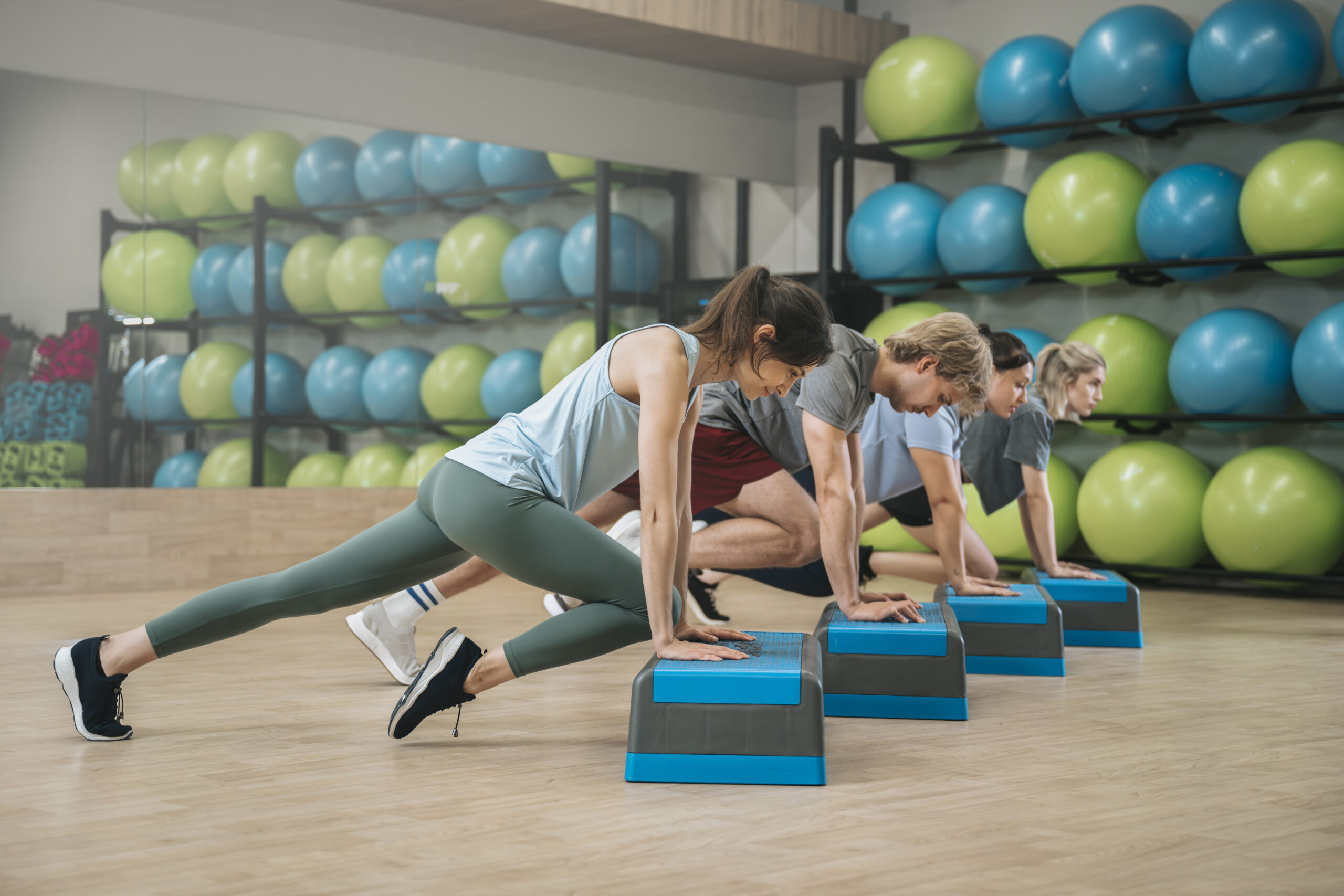 Grupa osób ćwicząca z użyciem stepów w sali fitness z piłkami gimnastycznymi w tle