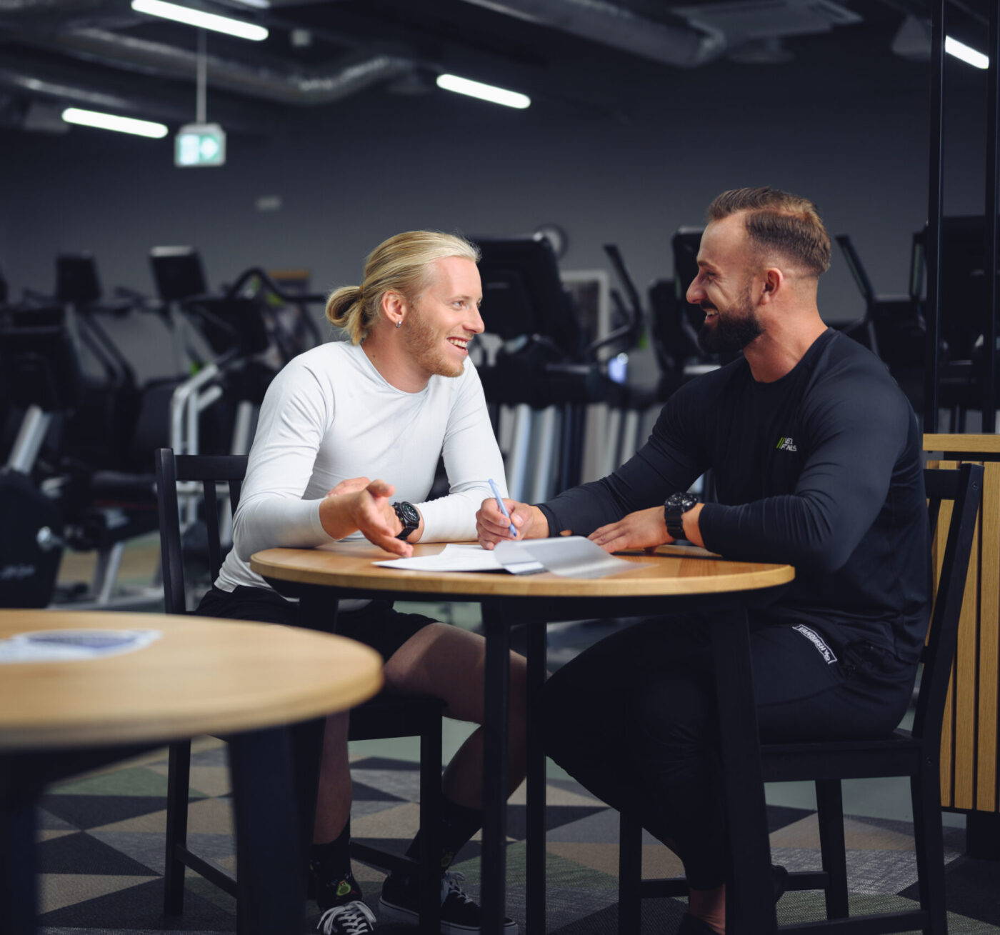 Trener z Well Fitness rozmawia z klientem przy okrągłym stoliku na siłowni, omawiając plan treningowy.