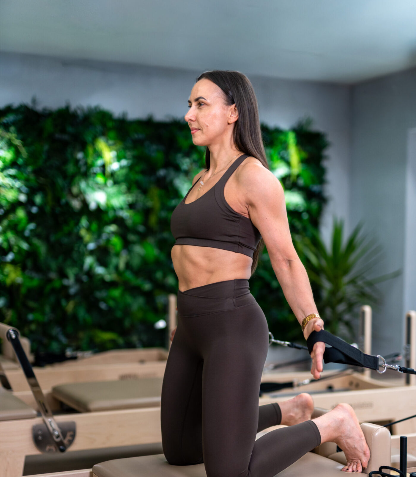 Kobieta wykonująca ćwiczenie pilates na reformerze , w studio z zieloną ścianą roślinną w tle