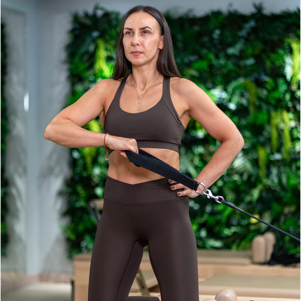 Kobieta wykonująca ćwiczenie pilates na reformerze , w studio z zieloną ścianą roślinną w tle