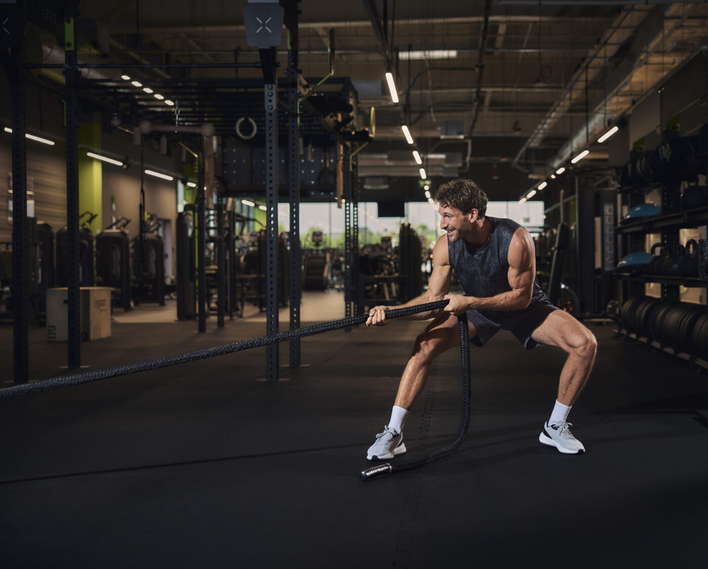 Mężczyzna w ciemnej koszulce bez rękawów wykonuje ćwiczenie z linami treningowymi w przestronnej siłowni.