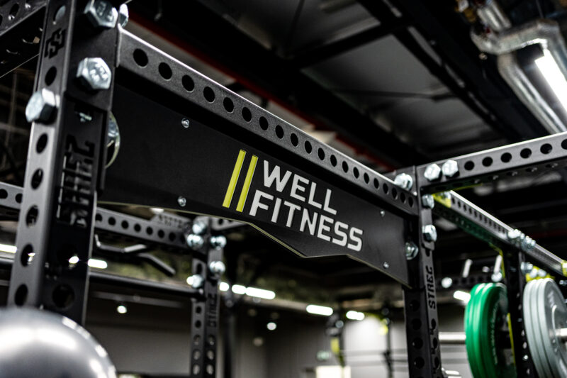 Detal czarnej stalowej konstrukcji treningowej z żółtym logotypem Well Fitness w strefie funkcjonalnej siłowni.