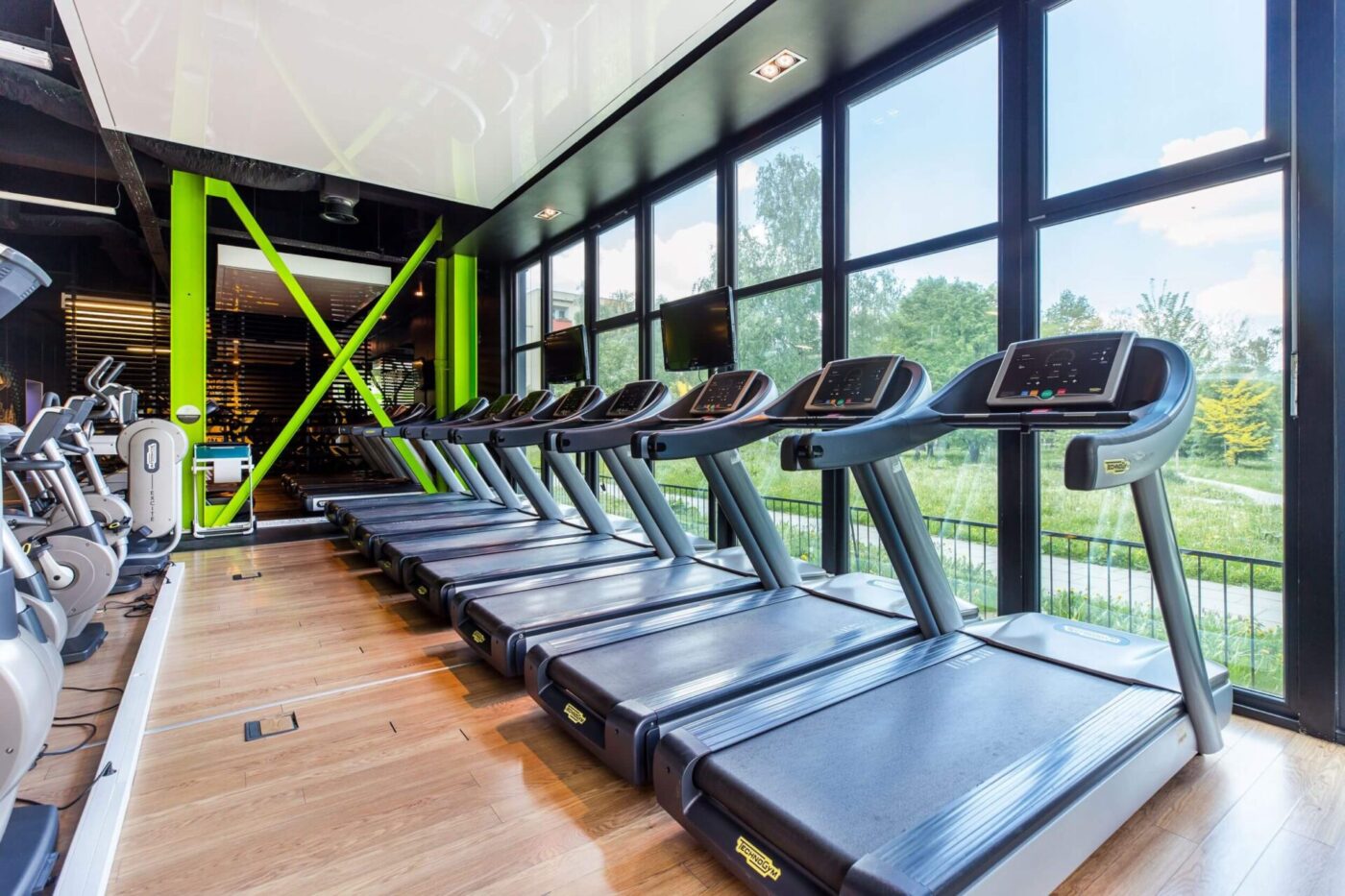 Nowoczesna strefa cardio w klubie Well Fitness Aleksandry w Krakowie z rzędem bieżni Technogym przy dużych przeszkleniach z widokiem na zieleń.