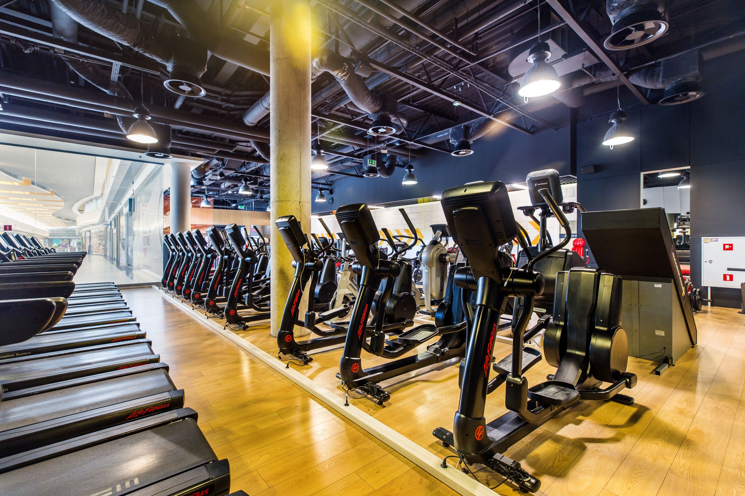 Strefa maszyn eliptycznych i cardio w Well Fitness Stawowa w Galerii Bronowice w Krakowie, nowoczesne oświetlenie i industrialny sufit.