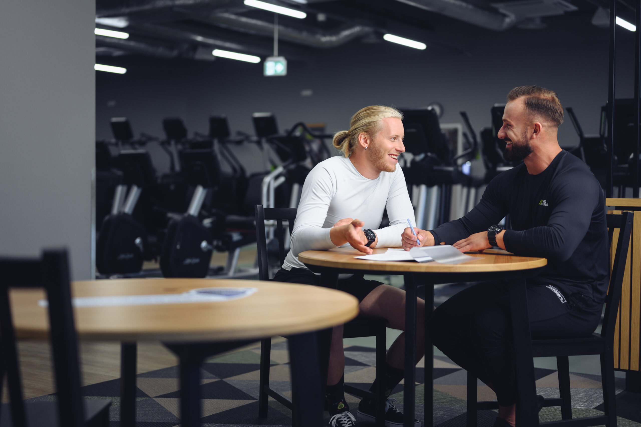 Two men during a consultation with a personal trainer at the Well Fitness gym - chatting at a table, cardio equipment in the background.