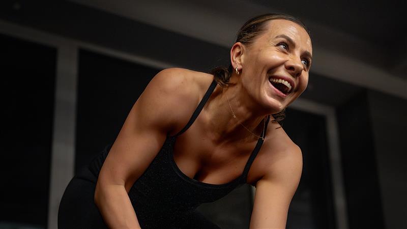 Uśmiechnięta kobieta w sportowym stroju podczas treningu w klubie fitness, patrząca z radością w bok.