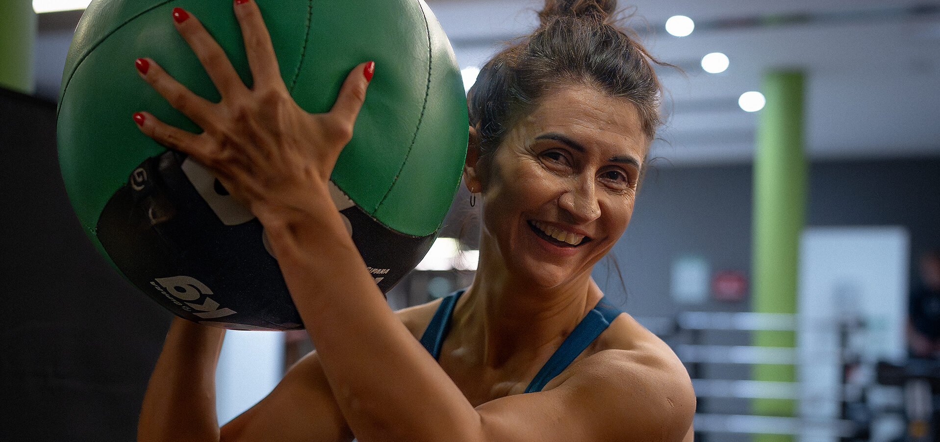 Uśmiechnięta, wysportowana kobieta w klubie fitness, trzymająca przy ramieniu dużą, zielono-czarną piłkę lekarską. Kobieta ma upięte włosy, jest ubrana w sportowy top i patrzy bezpośrednio w stronę obiektywu. W tle widoczna jest przestrzeń siłowni z maszynami do ćwiczeń i charakterystycznym zielonym filarem.
