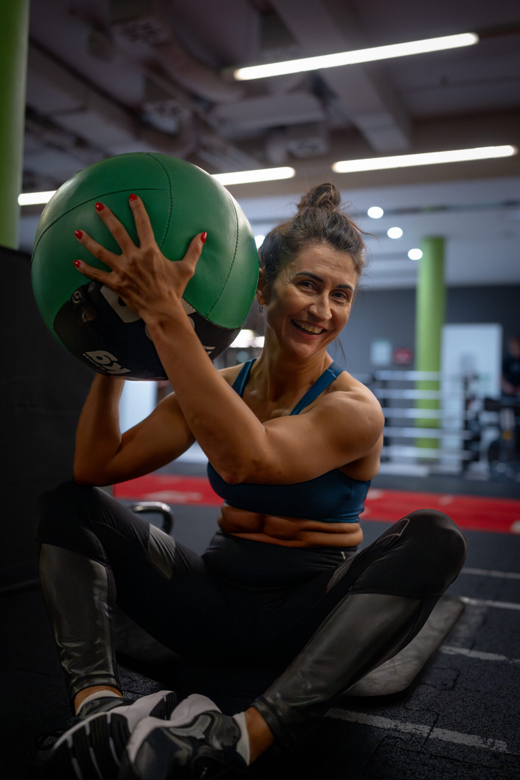 Uśmiechnięta, wysportowana kobieta w klubie fitness, trzymająca przy ramieniu dużą, zielono-czarną piłkę lekarską. Kobieta ma upięte włosy, jest ubrana w sportowy top i patrzy bezpośrednio w stronę obiektywu. W tle widoczna jest przestrzeń siłowni z maszynami do ćwiczeń i charakterystycznym zielonym filarem.