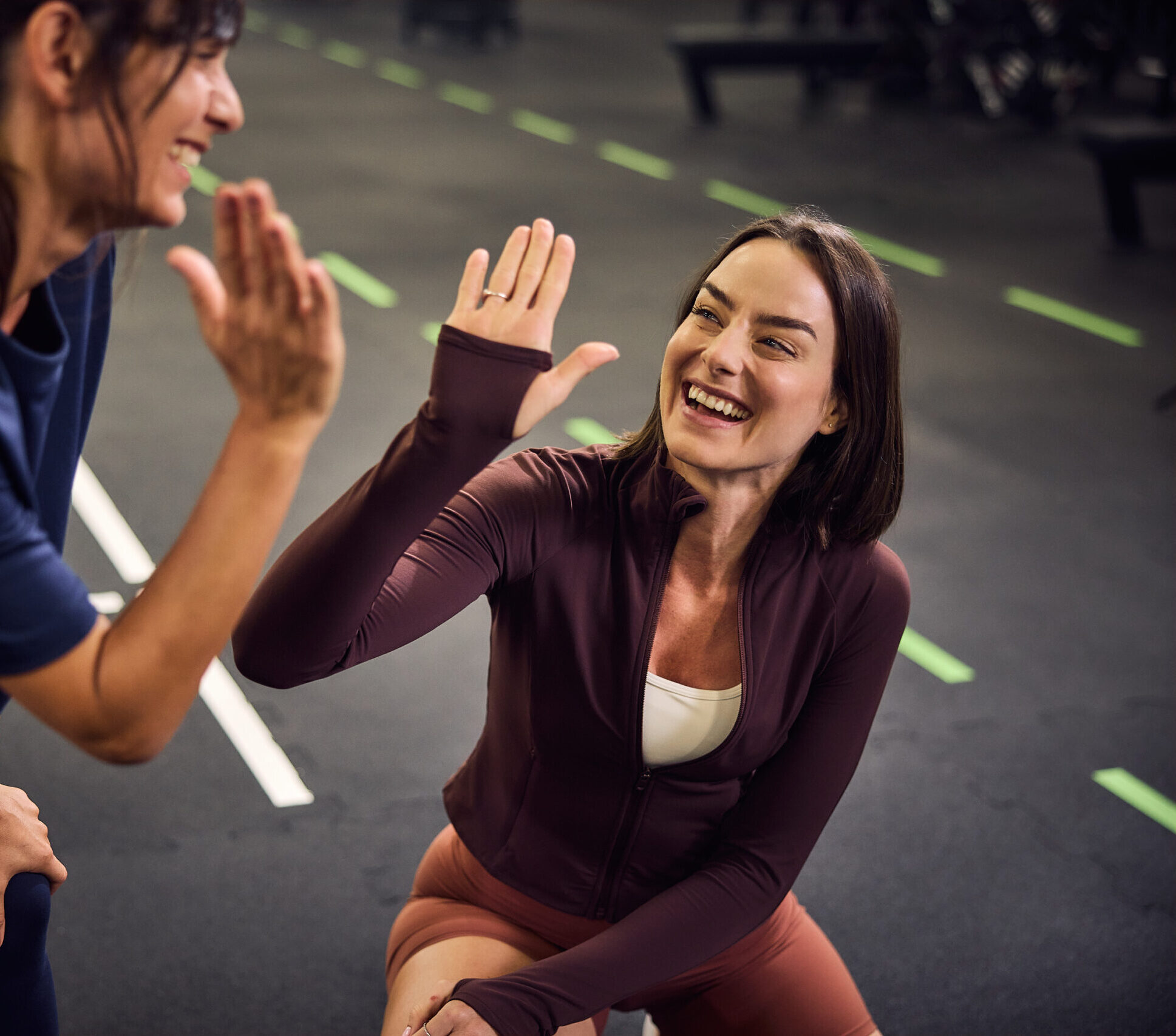 Dwie uśmiechnięte kobiety w strojach sportowych przybijają „piątkę” podczas treningu na siłowni. Jedna z nich klęczy na macie, a druga pochyla się w jej stronę, celebrując wspólnie wykonane ćwiczenie w nowoczesnym, sportowym wnętrzu.