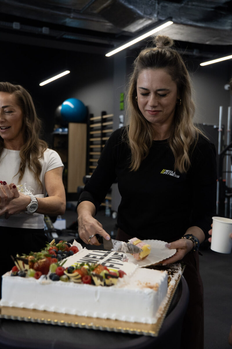 Kobieta w czarnej koszulce z logo Well Fitness kroi duży, prostokątny tort udekorowany świeżymi owocami (truskawkami, borówkami, malinami) podczas uroczystego otwarcia klubu. Na torcie widoczny napis "Well Fitness".