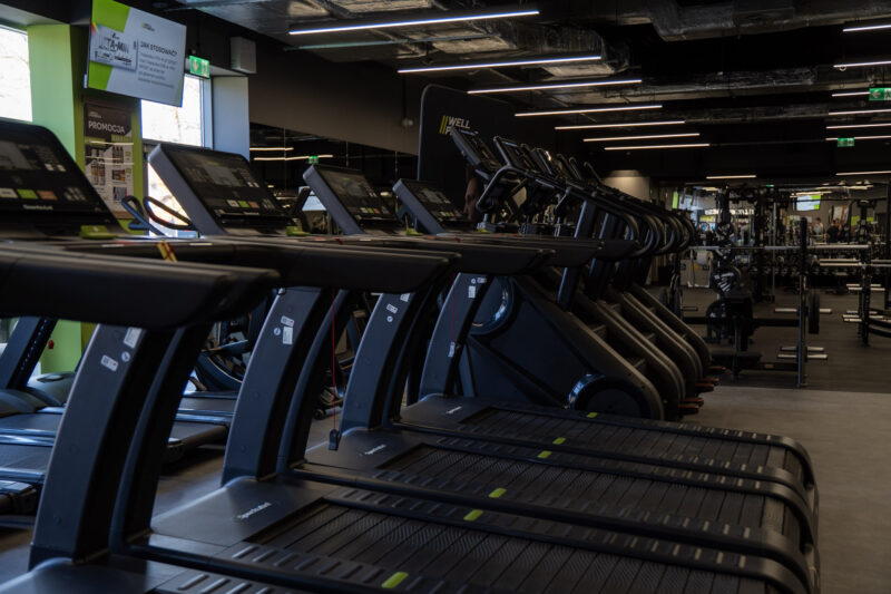Close-up of the row of modern treadmills at the Well Fitness club; the reception counter with the brand logo and the drinks fridge are visible in the background.