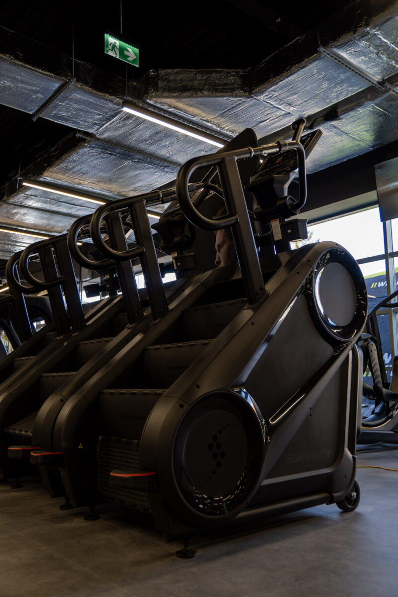 Construction details of the staircase-type cardio machines, visible against the bright window and industrial ceiling of the fitness club.