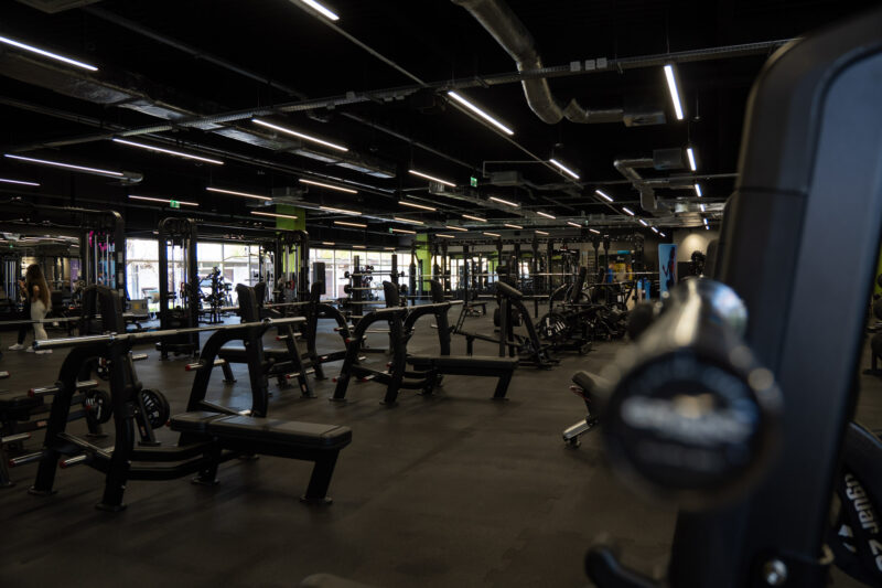 A wide shot of the free weights area with numerous exercise benches, griffins and dumbbells under long lines of ceiling lighting.