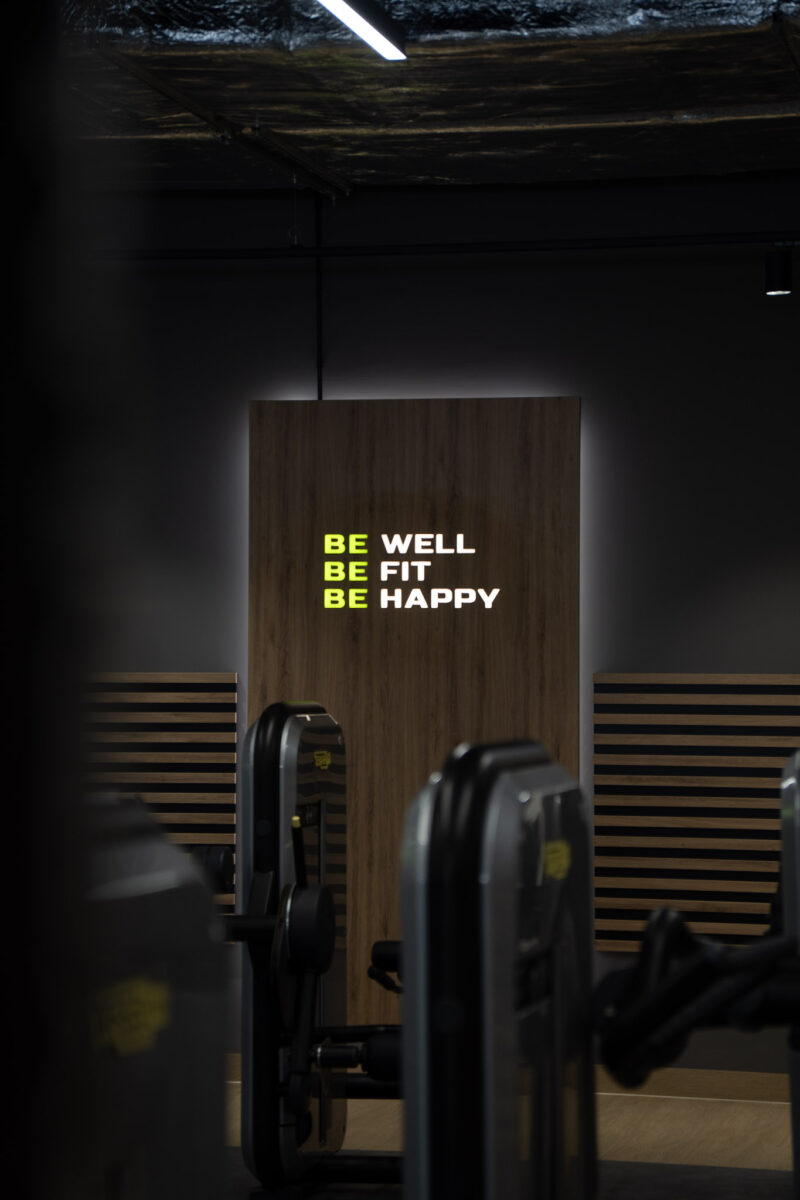 Illuminated motivational sign on wooden panel: „BE WELL, BE FIT, BE HAPPY” inside the Well Fitness Club.