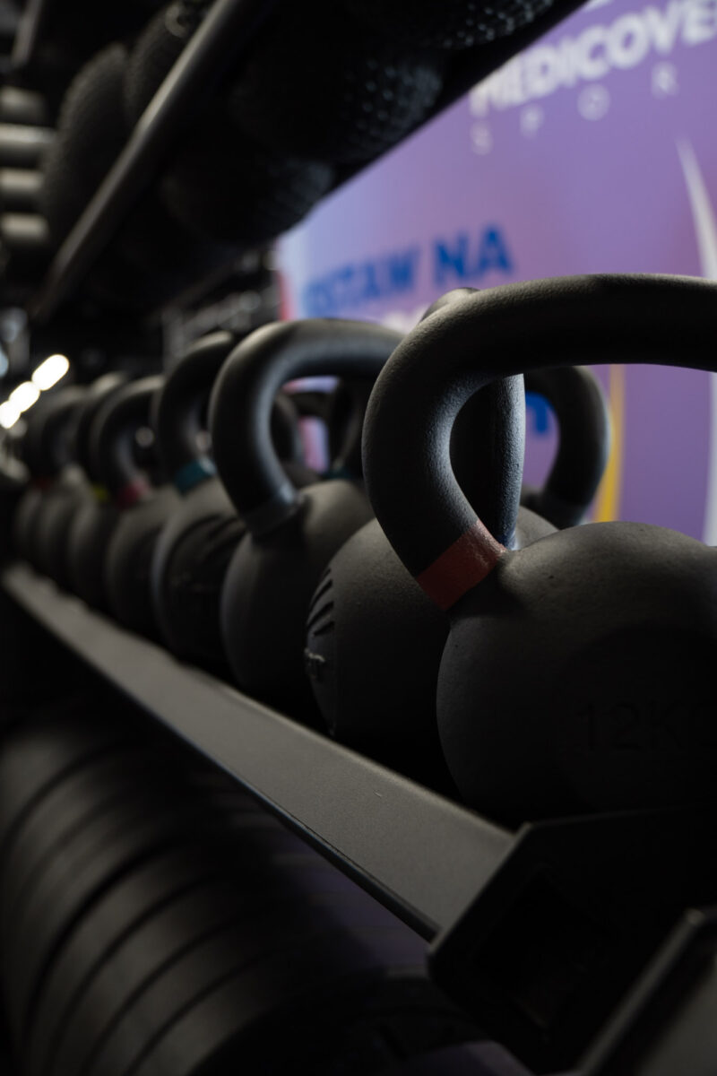 Rząd czarnych hantli kettlebell o różnej wadze ułożonych na metalowym regale w nowoczesnej siłowni. Na pierwszym planie zbliżenie na uchwyt kettlebella z czerwonym oznaczeniem, w tle widoczny fragment fioletowego baneru Medicover Sport.