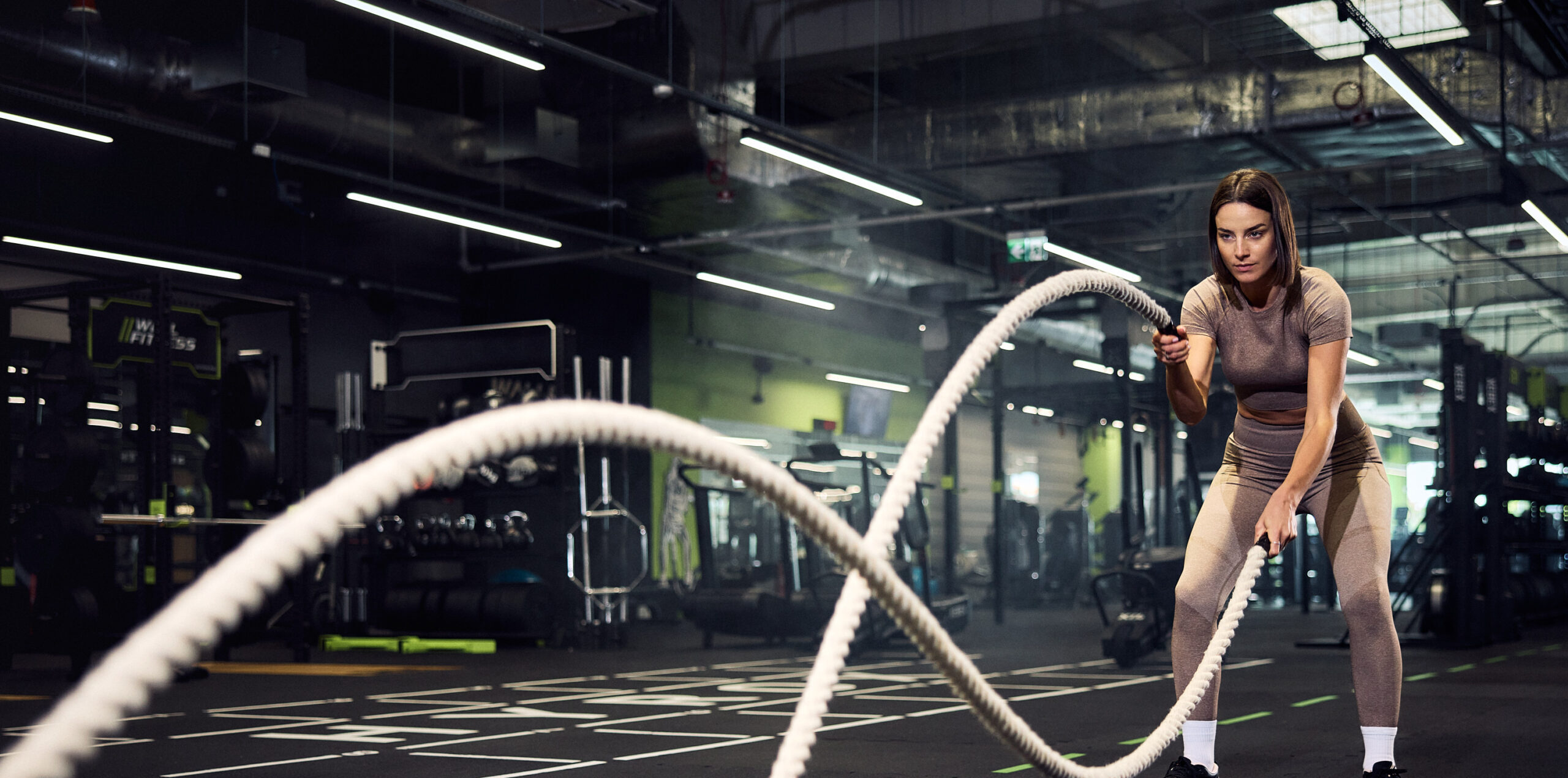 Kobieta trenująca z linami battle ropes w nowoczesnym wnętrzu siłowni Well Fitness.