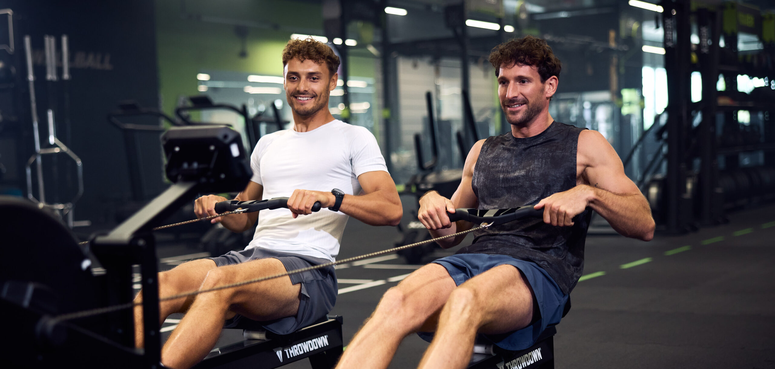 Dwaj mężczyźni z uśmiechem wykonujący trening na maszynach wioślarskich na siłowni, co ilustruje korzystanie z darmowych kalkulatorów fitness wspierających planowanie aktywności fizycznej.