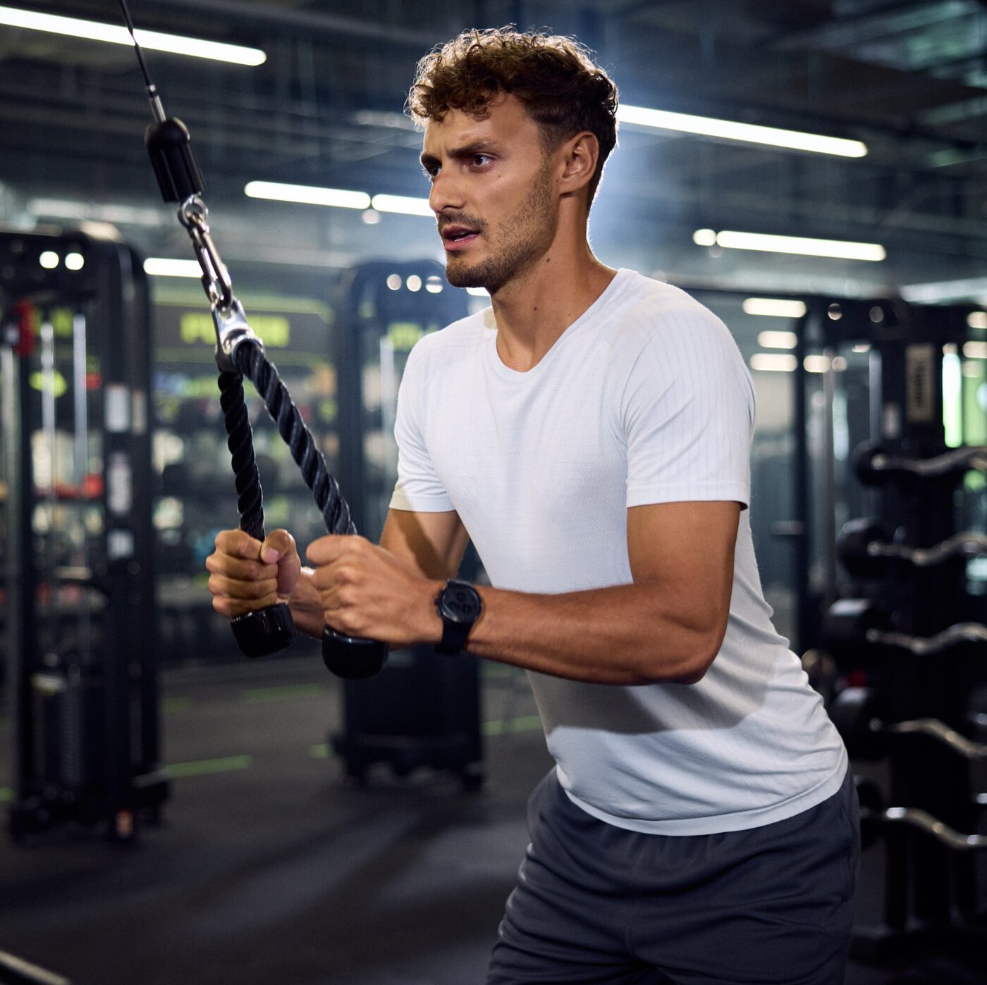 Młody mężczyzna w białej koszulce sportowej i ze zegarkiem na nadgarstku wykonuje prostowanie ramion na wyciągu górnym z liną w nowoczesnej siłowni.