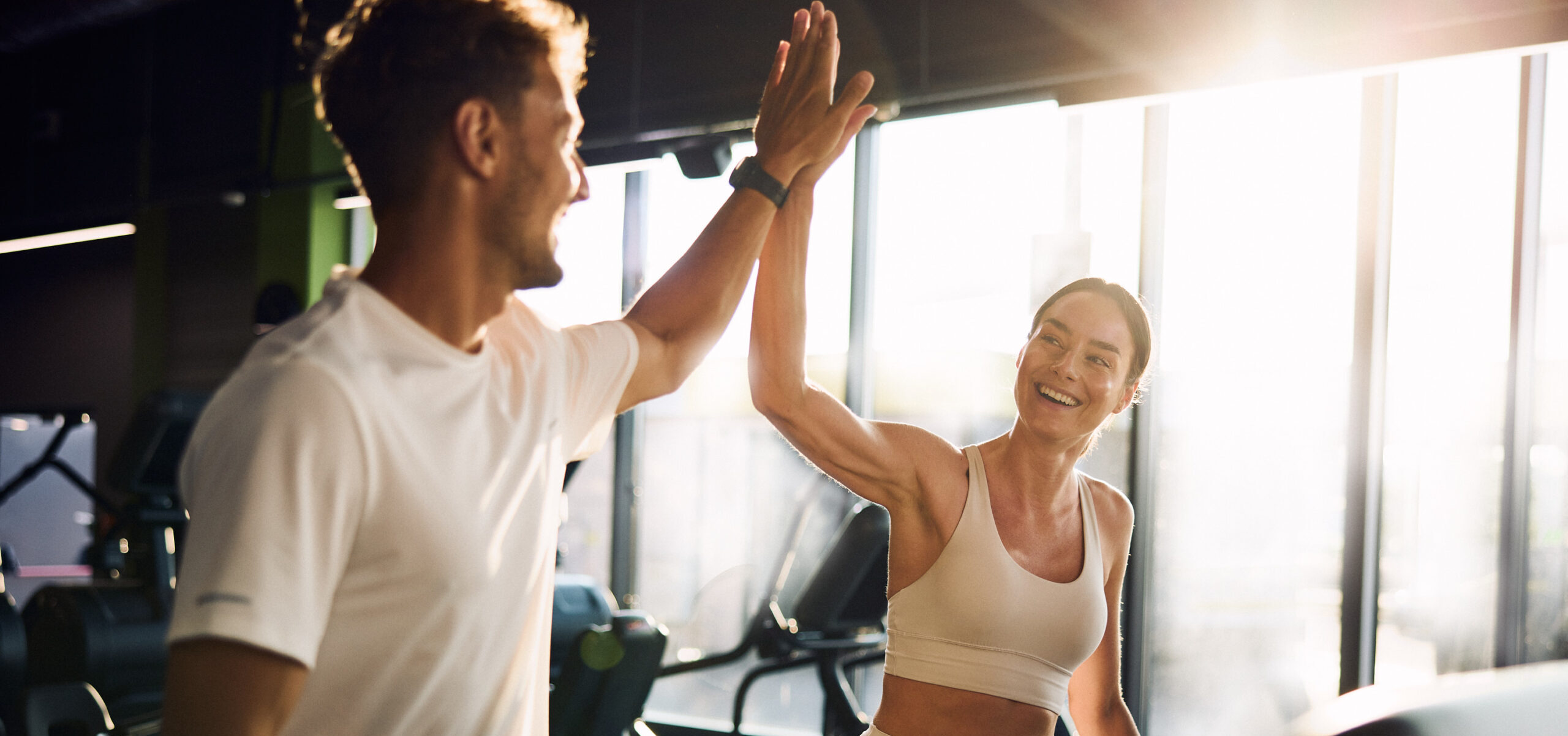 Mężczyzna i uśmiechnięta kobieta przybijają piątkę podczas treningu na siłowni Well Fitness.
